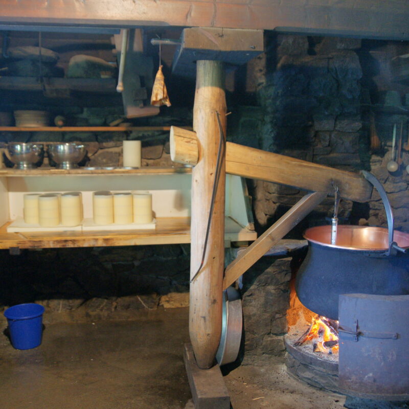 Calderone di rame tradizionale sul fuoco in un caseificio artigianale, con forme di formaggio in stagionatura sugli scaffali