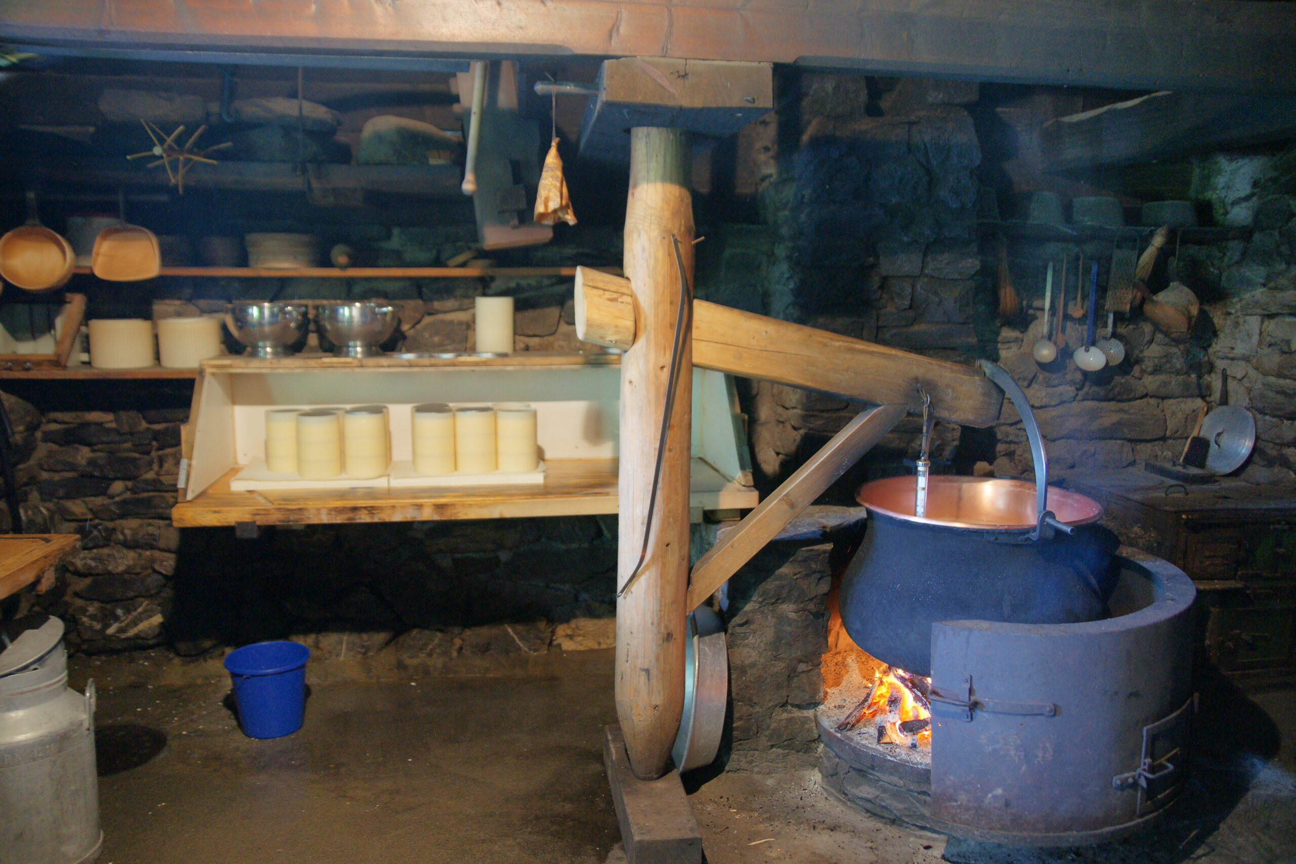 Calderone di rame tradizionale sul fuoco in un caseificio artigianale, con forme di formaggio in stagionatura sugli scaffali