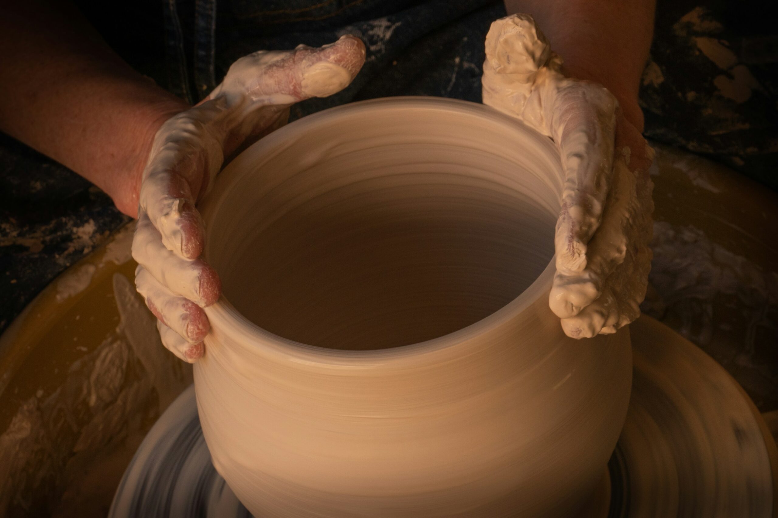 Primo piano di mani coperte di argilla che modellano un vaso su un tornio da ceramista in movimento