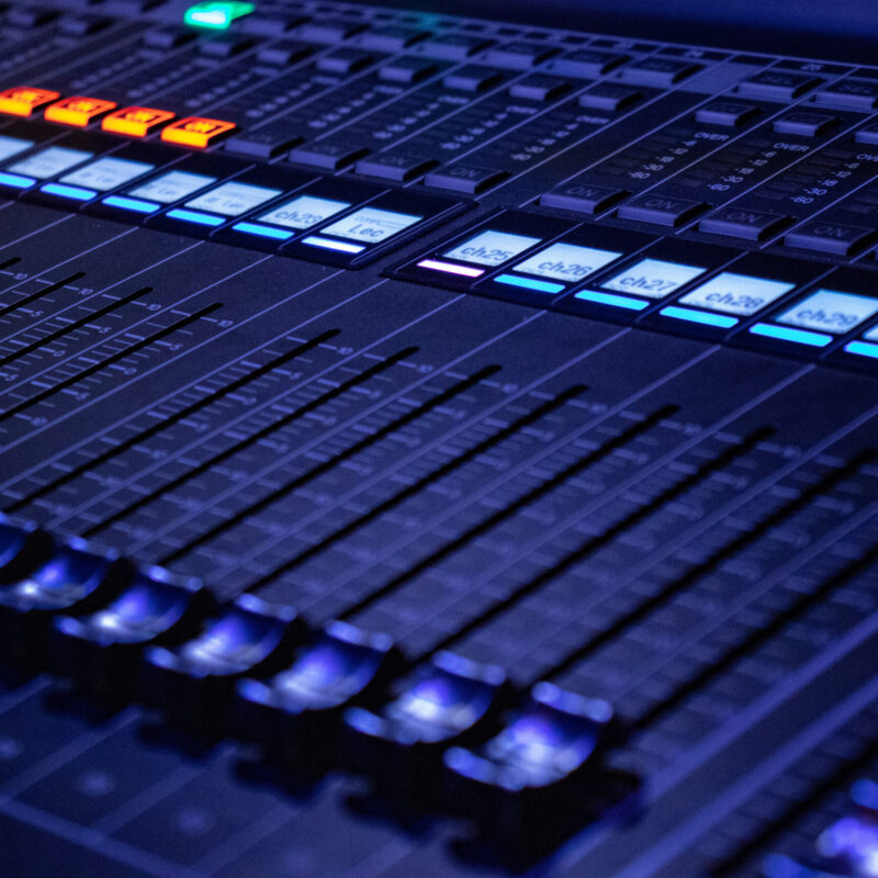 Primo piano di un mixer audio con cursori e pulsanti illuminati durante un evento live