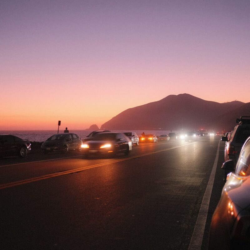 Fari di auto accesi al tramonto su strada costiera, guida con luce scarsa