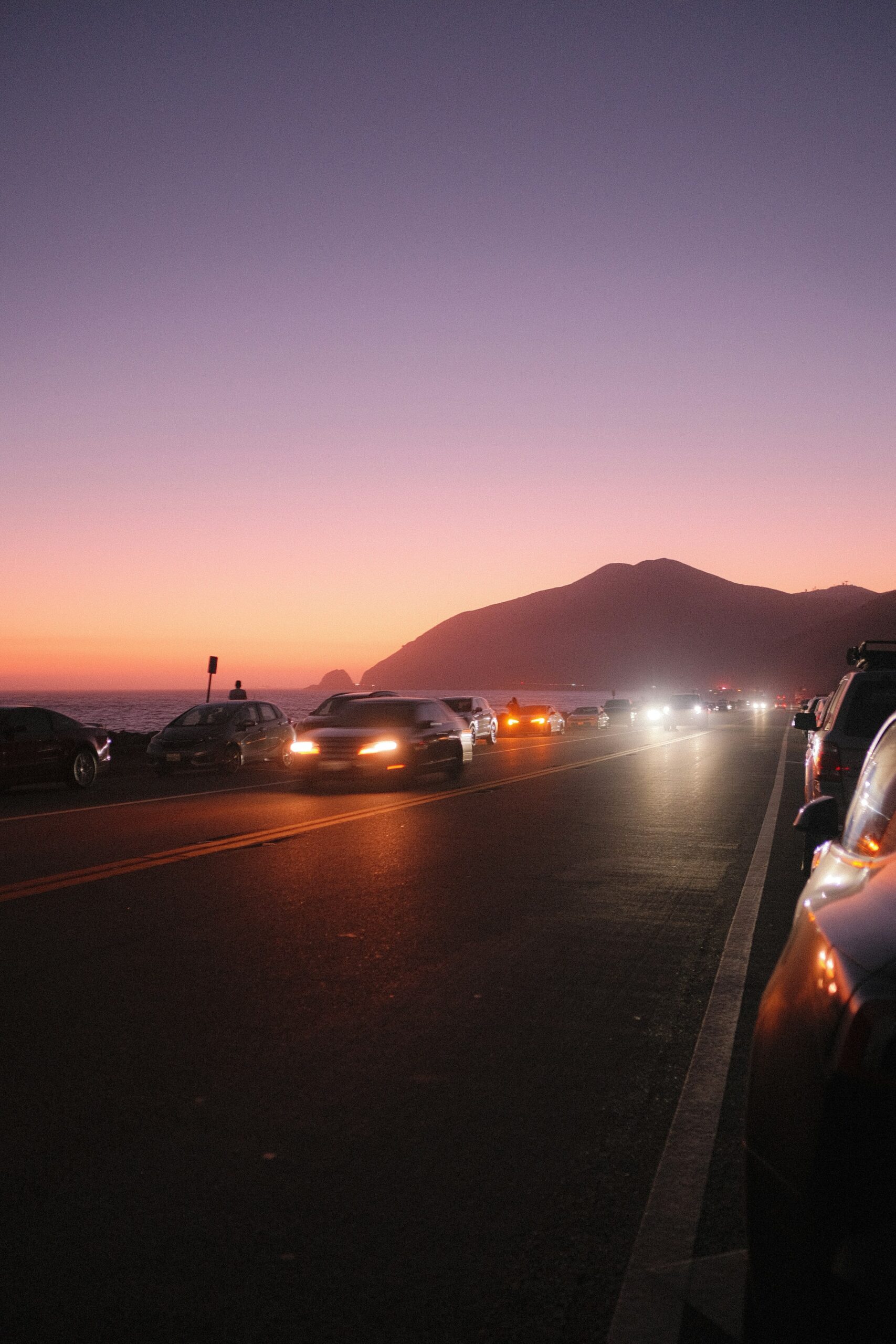 Fari di auto accesi al tramonto su strada costiera, guida con luce scarsa