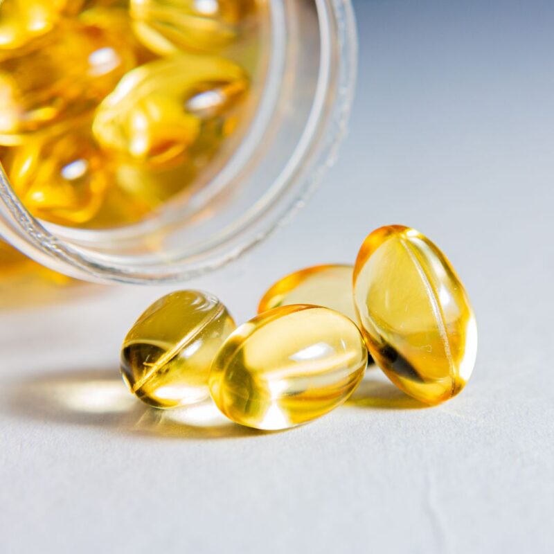 Capsule morbide dorate accanto a un contenitore trasparente su fondo chiaro, fotografia illustrativa di integratori