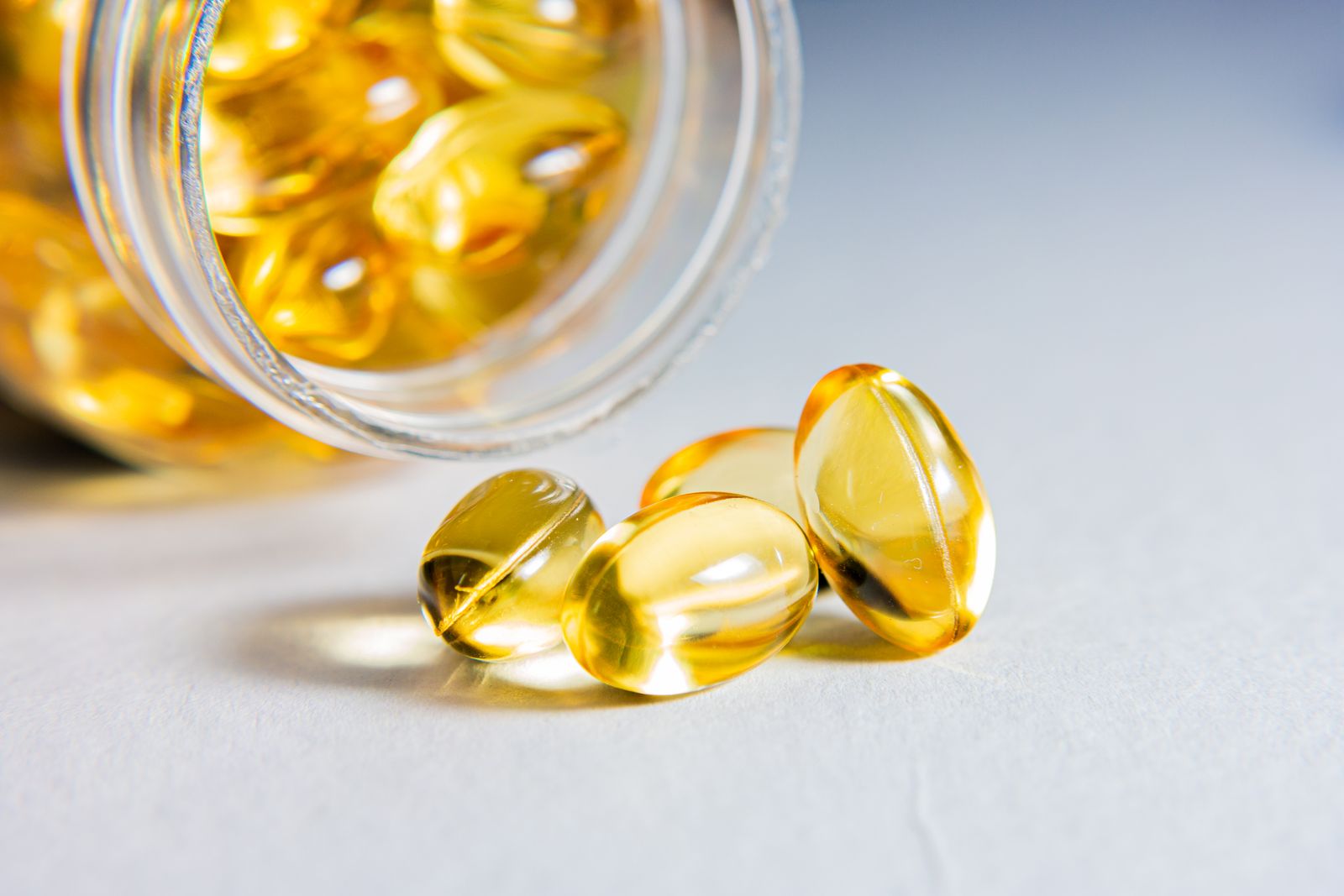 Capsule morbide dorate accanto a un contenitore trasparente su fondo chiaro, fotografia illustrativa di integratori